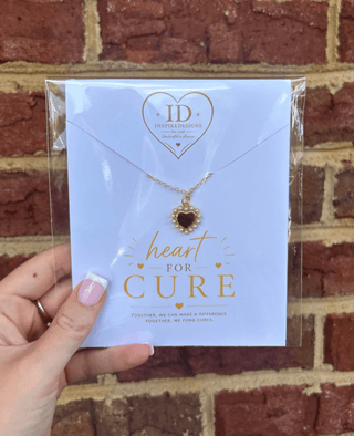 A gold heart pendant necklace with a dark red heart inlay, displayed on a light blue card that reads 'heart FOR CURE' with the ID Inspirations logo. The card is held by a hand against a red brick wall background.