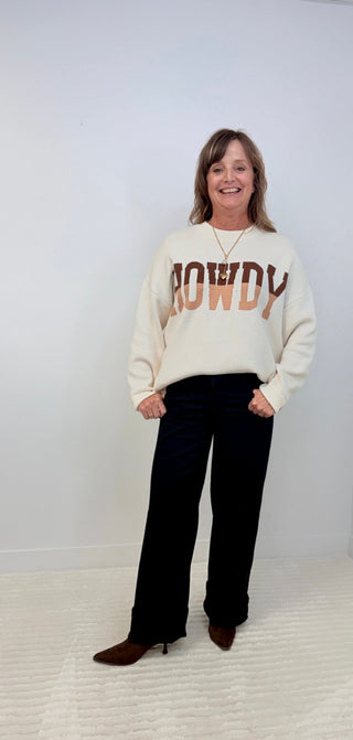 A woman modeling a cream-colored crewneck sweatshirt with 'HOWDY' printed across the chest in brown and orange, paired with black wide-leg pants and brown boots, standing against a plain white wall.