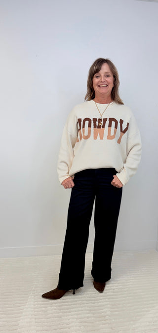 A woman modeling a cream-colored crewneck sweatshirt with 'COWDY' printed across the chest in brown and orange, paired with black wide-leg pants and brown boots, standing against a plain white wall.