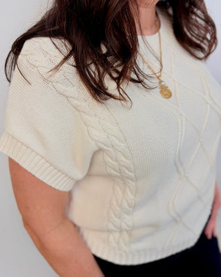 A close-up of a woman wearing a cream-colored short-sleeve cable-knit sweater, showing the detailed knit texture and diamond cable patterns.