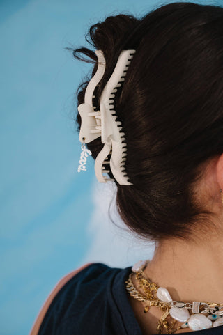 A close-up lifestyle shot of a person wearing a large, cream-colored claw hair clip to secure their dark hair. The person is shown from the back/side, wearing a navy top and layered gold and white necklaces, set against a solid light blue background.