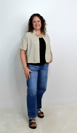 Woman wearing a beige cropped button down, black top, blue jeans, and sandals on a white background