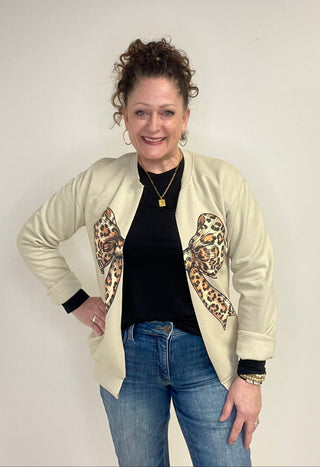 Woman wearing a cream sweatshirt cardigan with leopard print accents, black top, and blue jeans against a plain background.