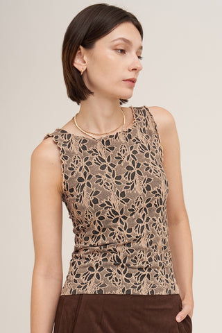 A woman wearing a sleeveless beige top with a black floral lace pattern, paired with brown bottoms, photographed against a plain light beige studio background.