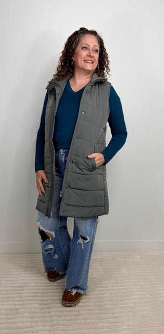 A woman modeling a long gray quilted puffer vest over a navy blue long-sleeve shirt and distressed blue jeans, standing against a plain light gray wall.