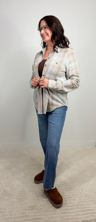 A woman modeling a light beige and white plaid button-down shirt over a dark brown top, paired with blue jeans and brown suede loafers, standing against a plain light gray wall.