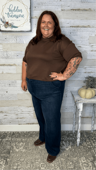 Woman wearing a brown shirt and blue jeans standing in front of a white wooden wall with a small table and pumpkins.