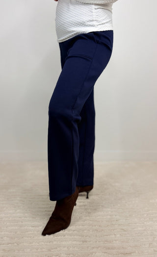 A person wearing navy blue straight-leg pants and brown pointed-toe heeled boots, standing on a light beige textured rug against a plain light gray wall.