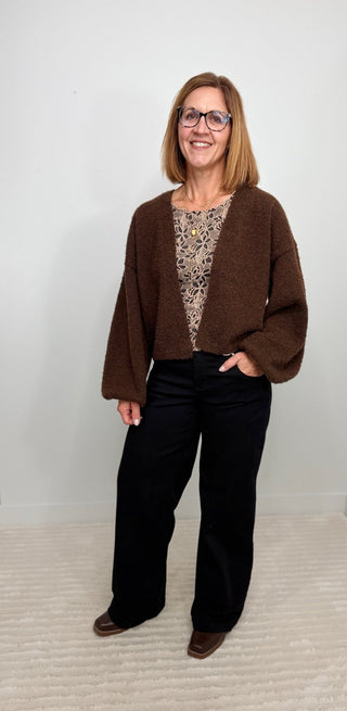 A woman modeling a brown fuzzy cropped cardigan over a patterned top and black pants, standing against a plain light gray wall and textured cream carpet.