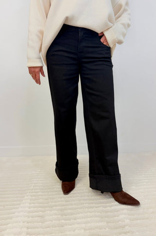 A person wearing black wide-leg cropped pants with rolled cuffs, paired with a cream sweater and brown pointed-toe flats, standing against a white wall and textured white floor.