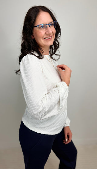 A woman with dark hair and blue-framed glasses wearing a white textured long-sleeve blouse and dark navy pants, posing against a neutral light gray studio background.