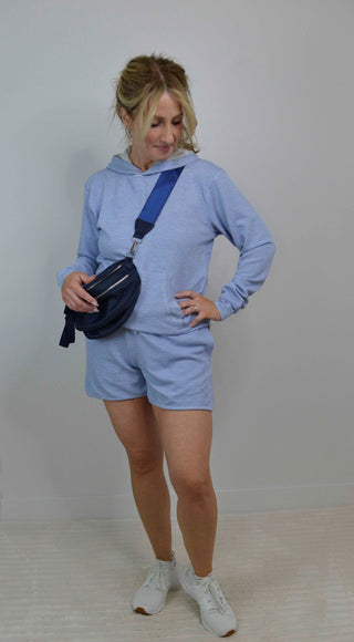 Woman wearing a light blue hoodie and shorts set with a navy fanny pack against a plain background