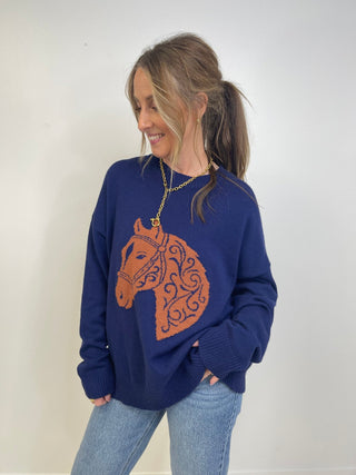 A woman wearing a navy blue knit sweater with a large orange horse head graphic on the front, paired with light blue jeans and a gold chain necklace, standing against a plain white wall.