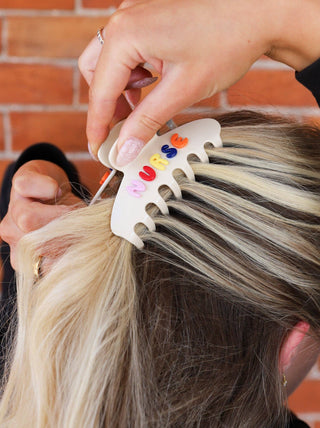 Person using a hair color comb with colorful letters "nurse" to dye hair, against a brick wall background.