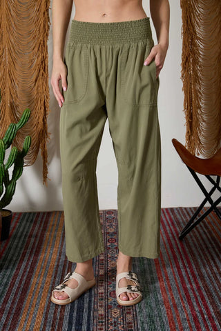 Person wearing olive green wide-leg pants standing in a room with a plant and chair.