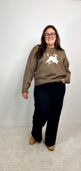 A woman wearing a brown knit sweater with a white and tan dog graphic on the chest, paired with black pants and tan boots, standing against a plain light gray wall.