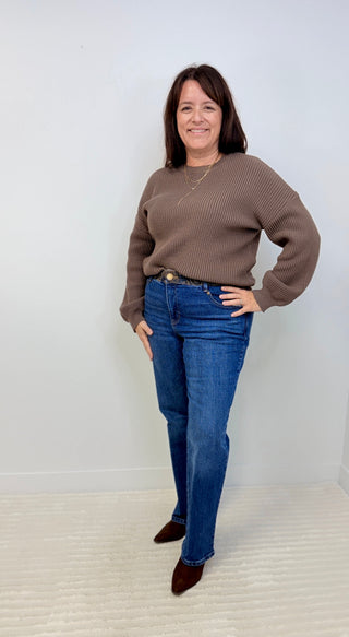 A woman modeling a brown ribbed knit sweater and blue jeans against a plain white wall and light carpet.