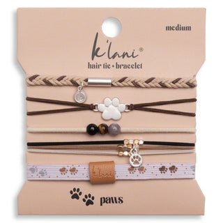 A set of pet hair tie and bracelet accessories displayed on a beige branded backing card. The set includes braided and elastic bands in tan, brown, black, and pale pink, with decorative charms such as a silver paw print, small beads, and a faux-leather tab.