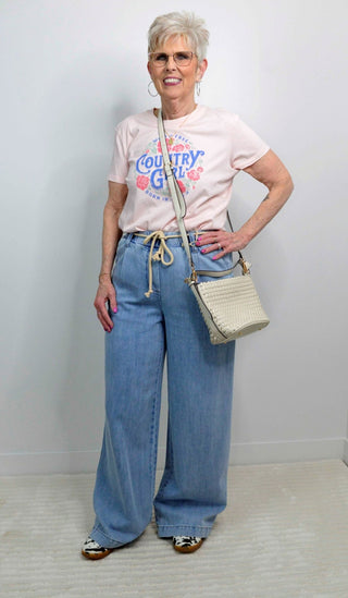 Woman wearing a 'Country Girl' t-shirt with wide-leg jeans and a beige handbag against a white background