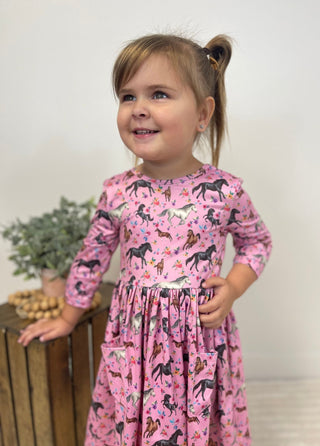 A young girl with a high ponytail smiling and looking to the side, wearing a pink long-sleeve dress with a black and multicolor horse pattern. She is standing next to a wooden crate with a small potted plant against a plain white wall.