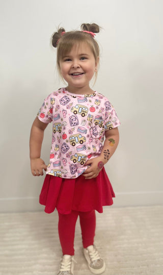A young girl with her hair in two buns stands smiling, wearing a pink short-sleeve t-shirt with a colorful school bus pattern, a red pleated skirt, red leggings, and white sneakers.