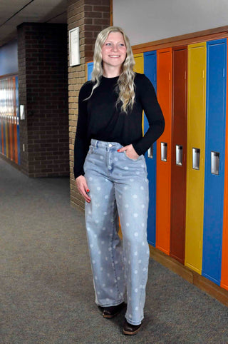A woman stands in a school hallway wearing a black long-sleeve top and light blue jeans with white polka dots, with colorful lockers behind her.
