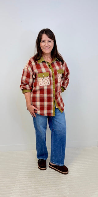 A woman modeling a red and green plaid button-up shirt with patchwork pockets, paired with blue jeans and dark brown slip-on shoes, standing against a plain light gray wall.