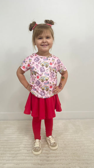 A young girl with brown pigtails stands with hands on hips, wearing a pink short-sleeve t-shirt with a colorful dump truck pattern, a red pleated skirt, red leggings, and beige sneakers, set against a plain white wall and light carpet.