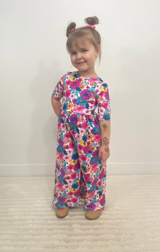 A toddler girl with her hair in two buns stands on a light beige carpeted floor against a plain off-white wall, wearing a short-sleeve wide-leg jumpsuit with a vibrant multicolor floral pattern.