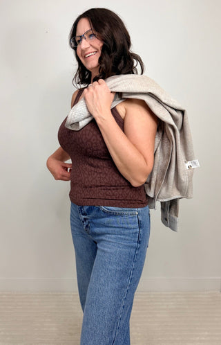 A woman wearing a brown patterned tank top and blue jeans stands indoors, holding a light gray knit wrap or blanket over her shoulder.