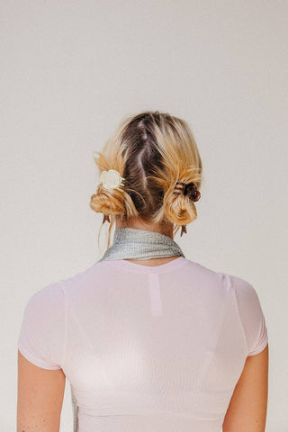 A woman viewed from behind wearing a pale pink ribbed short-sleeve top and a metallic silver neck scarf, with blonde hair styled in a low bun with a white flower accessory.
