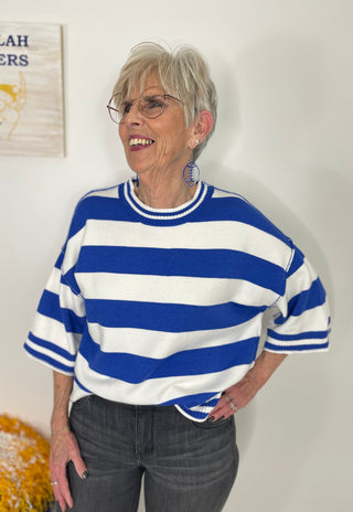 An older woman with short gray hair and glasses is smiling while wearing a blue and white horizontally striped knit sweater with short sleeves and gray jeans, standing against a white wall.