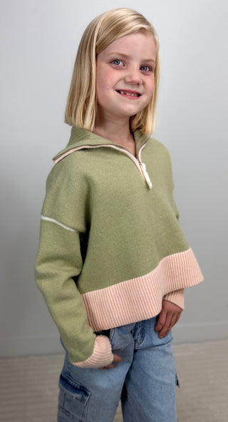 A young blonde girl wearing a sage green half-zip sweater with a light pink ribbed hem and cuffs, paired with blue jeans, standing against a neutral gray background.