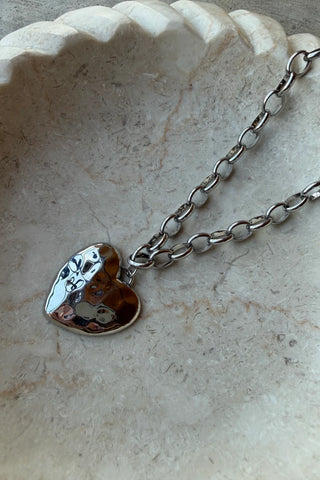 A silver heart-shaped pendant necklace with a hammered finish and a small white enamel heart, resting on a beige stone dish.