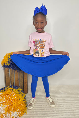 A young girl stands holding out a bright royal blue skirt, wearing a light pink graphic T-shirt, matching blue leggings, and a large blue bow in her hair. She is posed in front of a plain light wall with decorative pom-poms on the left.