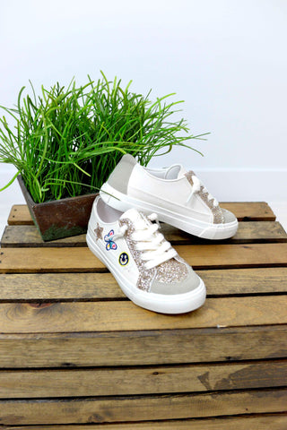 A pair of low-top children's sneakers in white and glittery silver, displayed on a wooden crate next to a potted green plant against a white wall.