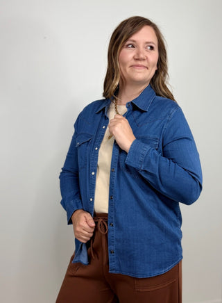 A woman wearing a blue denim button-up shirt over a cream top and brown pants, standing against a plain light gray wall.