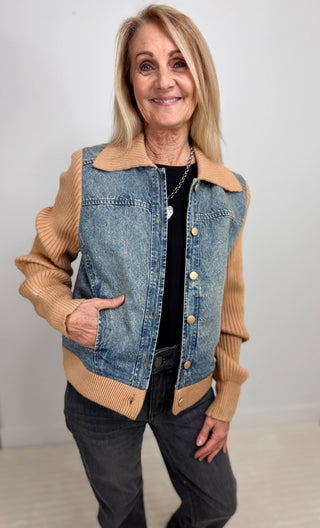 A smiling blonde woman wearing a unique hybrid jacket with a blue denim body and tan ribbed knit sleeves and collar, paired with a black top and dark jeans.