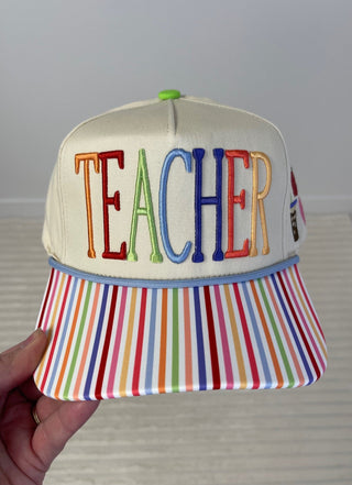 Cap with 'TEACHER' in colorful letters on a striped brim, held against a neutral background.