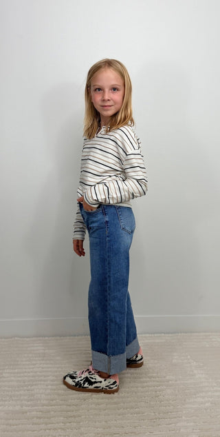 A young girl with blonde hair stands in a room wearing a long-sleeve white shirt with thin black stripes, blue jeans, and black-and-white patterned shoes.