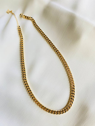 A gold-toned chain necklace with a textured curb-style link pattern, displayed on a soft, off-white fabric background.