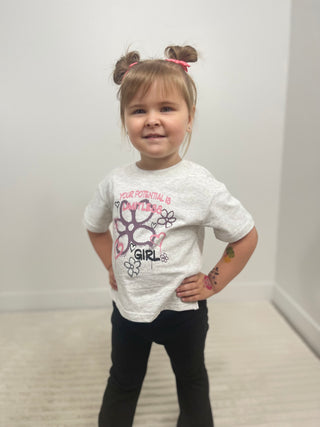 A young girl with her hair in two buns stands with hands on hips wearing a light gray graphic t-shirt that reads 'YOUR POTENTIAL IS LIMITLESS GIRL' with a pink and purple paw print design, paired with black pants.