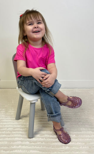 Child sitting on a small chair wearing a pink shirt and purple jelly shoes.