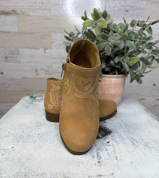 Pair of tan ankle boots for toddlers with decorative stitching on a rustic surface with a plant in the background.