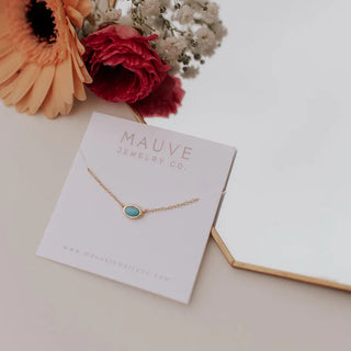 Gold necklace with a blue pendant on a Mauve Jewelry Co. card with flowers in the background.