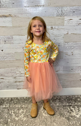 Young girl wearing a floral top and peach skirt standing against a wooden paneled wall.