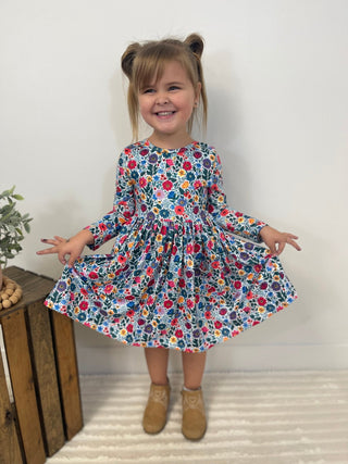 A young girl with her hair in two buns stands smiling and twirling in a long-sleeve dress with a colorful floral print. She is wearing tan suede boots and standing on a white textured rug in front of a plain white wall, with a rustic wooden crate and plant visible on the left.