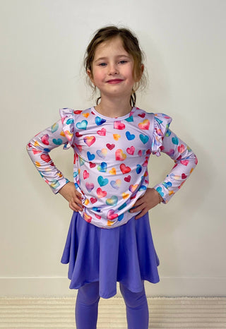Young girl wearing a colorful heart-patterned shirt and purple skirt against a plain background