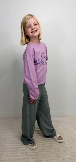 A young girl with blonde hair stands smiling, wearing a lavender long-sleeve crewneck sweatshirt with a smiley face graphic and dark gray wide-leg sweatpants.