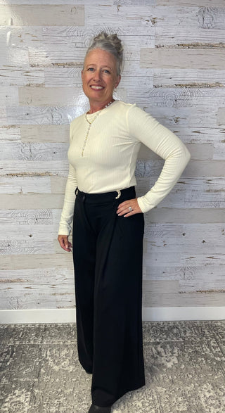 Woman wearing a white long-sleeve top and black pants against a textured wall.